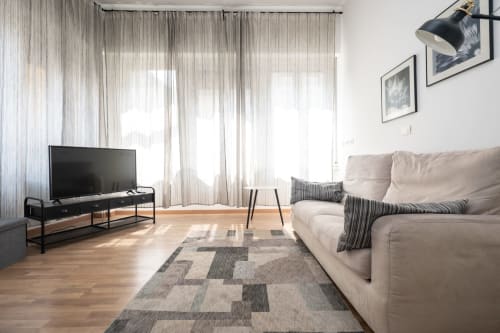 Soggiorno moderno a Milano con divano beige, TV, tappeto decorativo e luce naturale dalle tende trasparenti
