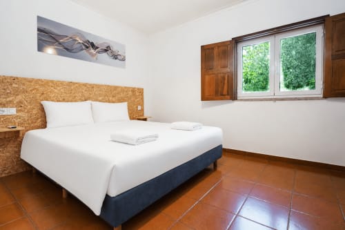 Quarto com roupa de cama branca, cabeceira de cortiça, piso de azulejo terracota e janela com vista para o jardim na Quinta de Carvalhal em Guimarães