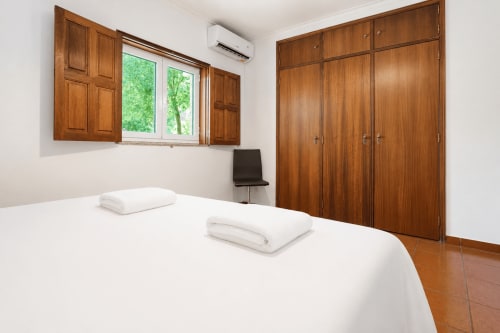 Quarto luminoso com cama branca, roupeiros de madeira, janela com vista para o jardim e piso de azulejo terra cotta na Quinta de Carvalhal