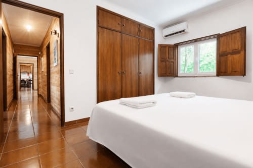 Quarto espaçoso na Quinta de Carvalhal com cama branca, guarda-roupas de madeira, janelas com vista para o jardim e pisos em terracota