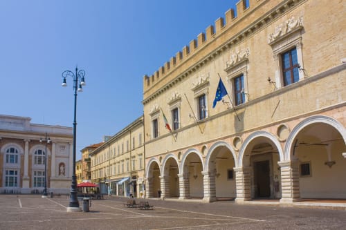Palazzo rinascimentale in piazza italiana con porticato al piano terra, bandiere e cielo azzurro limpido