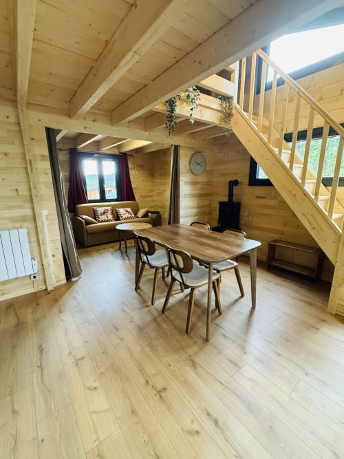 Intérieur de lodge en bois avec table à manger, canapé marron, cheminée et escalier vers la mezzanine aux Cabanes du Belvédère, Serrières-sur-Ain