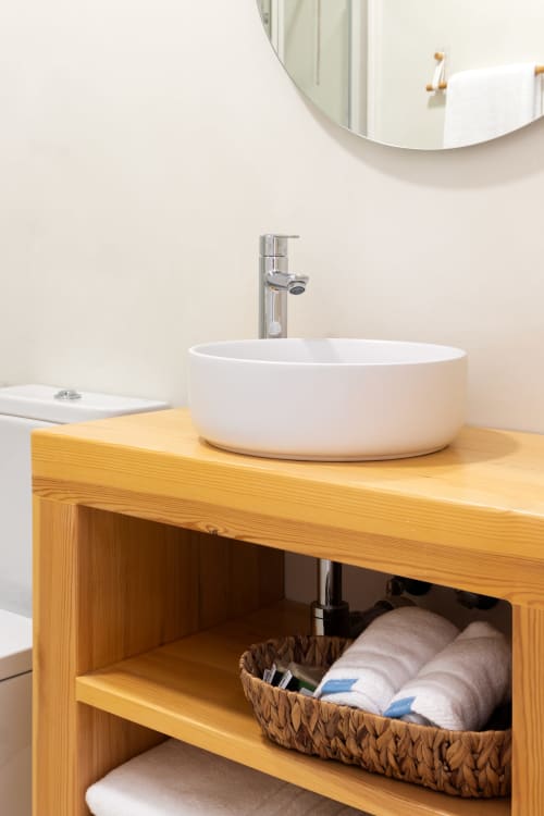 Elegante tocador de baño con lavamanos de cerámica blanca, mueble de almacenamiento de madera y espejo redondo en el hotel Ona vacaciones de Gandía