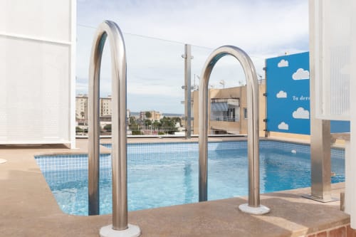 Moderna zona de piscina con duchas de arcos metálicos curvos en apartamento Ona vacaciones Gandía con solárium y vistas a la ciudad