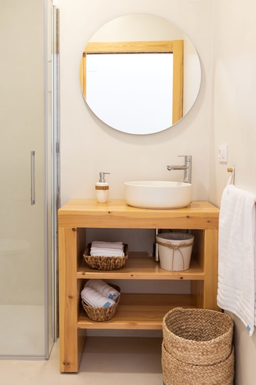 Moderno lavabo de baño con mueble de madera, espejo redondo y cestas tejidas en Ona vacaciones, Gandía