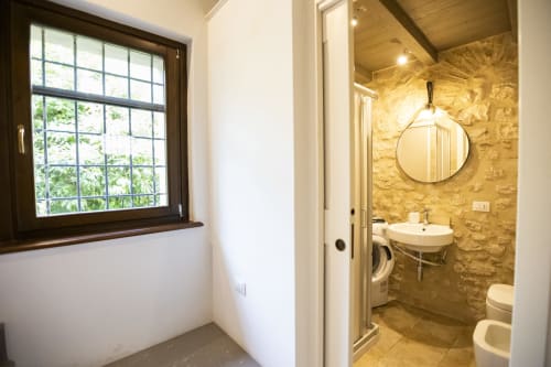 Bagno moderno a Le Mura Rooms a Norcia con muri in pietra, specchio rotondo, lavabo, lavatrice e vista dalla finestra
