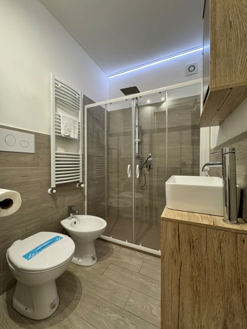 Bagno moderno nella stanza Cleopatra dell'Hotel Queen a Campofilone, con doccia in vetro, lavabo bianco, bidet e finiture in legno