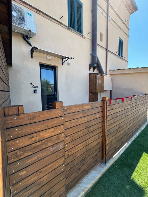 Vista giardino dell'Hotel Queen a Campofilone con staccionata in legno, sedie rosse e edificio beige con unità AC