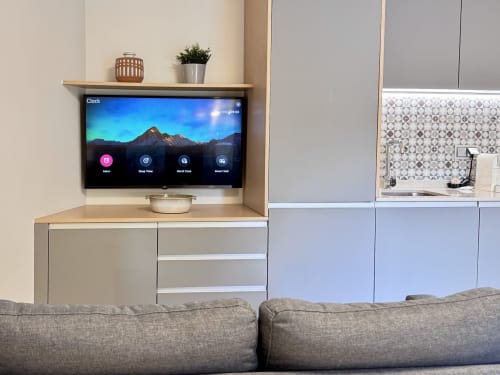 Salon moderne avec canapé gris, téléviseur mural affichant horloge, étagères bois et kitchenette à RIA CENTRUM