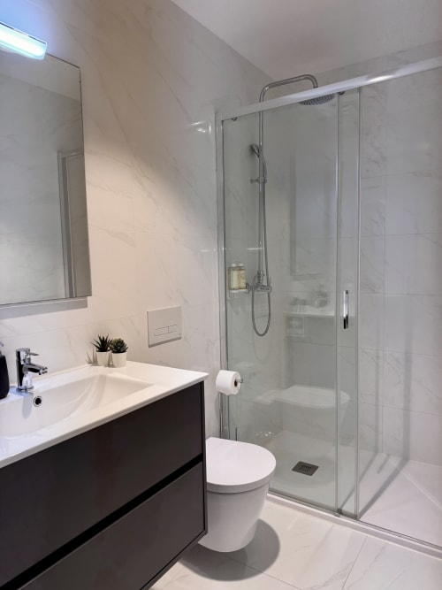 Salle de bain moderne à RIA CENTRUM Aveiro avec douche vitrée, lavabo blanc, meuble-lavabo sombre, toilettes et carrelage effet marbre