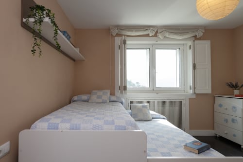 Dormitorio luminoso con paredes color melocotón, cama blanca y vistas a las montañas desde las puertas del balcón de la cabaña Vilaboa en Pontevedra