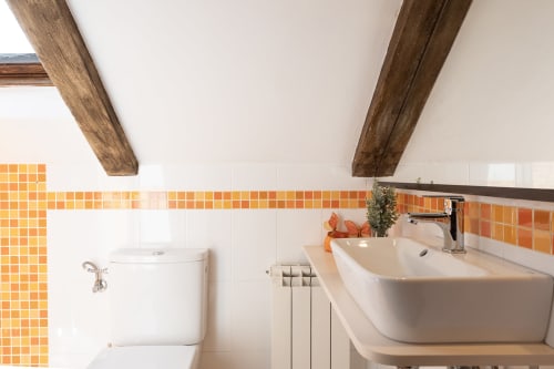 Baño moderno en casita Vilaboa con azulejos de mosaico naranja, lavamanos blanco, inodoro, vigas de madera y vistas a la montaña