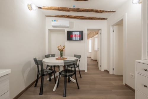 Zona pranzo moderna nell'appartamento di Cagliari con tavolo rotondo bianco, sedie nere, travi in legno e Netflix sulla TV a parete