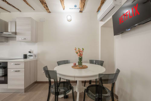 Zona cucina-soggiorno moderna nell'appartamento di Cagliari con tavolo rotondo bianco, sedie nere, travi in legno a vista e TV Netflix