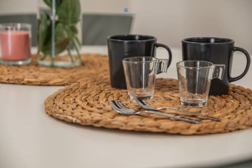 Tavolo apparecchiato con tazze nere, bicchieri trasparenti e cucchiai su tovaglietta intrecciata presso l'appartamento House Live Express a Cagliari