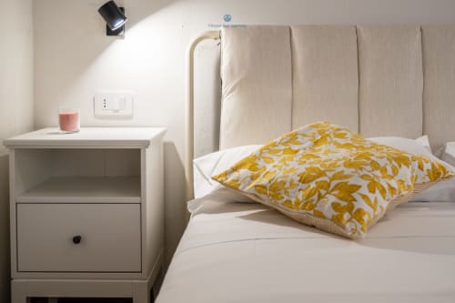 Accogliente camera da letto presso House Live Express a Cagliari con testata beige, comodino bianco, cuscino giallo con motivi e luce naturale