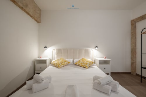Camera da letto moderna presso House Live Express Cagliari con letto bianco, cuscini gialli, comodini e asciugamani arrotolati