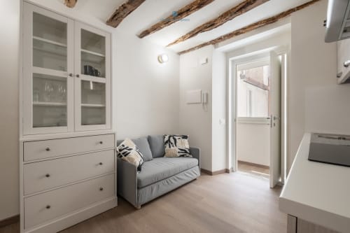 Zona living moderna con divano grigio, armadi bianchi, travi a vista e angolo cottura presso House Live Express