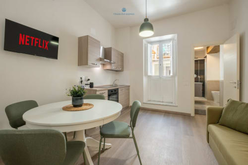 Cucina e zona pranzo moderna a Cagliari con TV Netflix, tavolo rotondo, sedie verdi e balcone con vista sulla città
