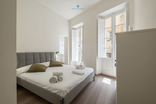 Camera moderna a House Live Express a Cagliari con letto imbottito grigio, biancheria bianca, pavimenti in legno e porte-finestre sul balcone