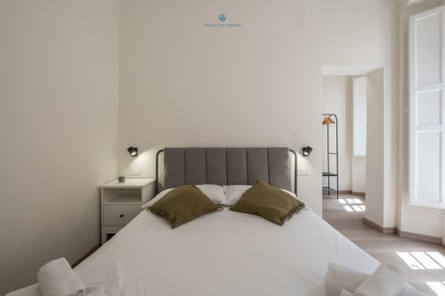 Camera da letto moderna e luminosa con testata imbottita grigia, biancheria bianca e cuscini verde oliva presso House Live Express Cagliari