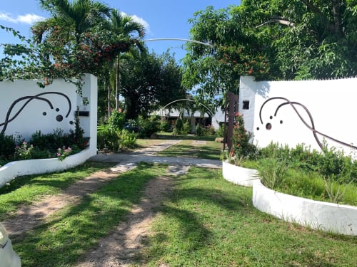 Lush tropical garden entrance at El Litoral Wellness Retreat with white walls, palm trees, and artistic sculptures.