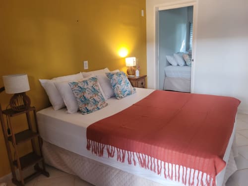Suite luminosa com paredes douradas, cama queen com manta vermelha, almofadas brancas, almofadas com estampa tropical e banheiro ensuite