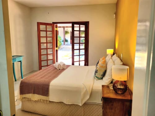 Quarto ensolarado na Casa do Sol Ilhabela com cama queen, portas de madeira abrindo para varanda com vista da piscina e parede de destaque em tons quentes de amarelo