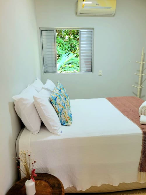 Quarto luminoso da Casa do Sol Ilhabela com almofadas brancas, almofada estampada azul, janela com vista para jardim exuberante