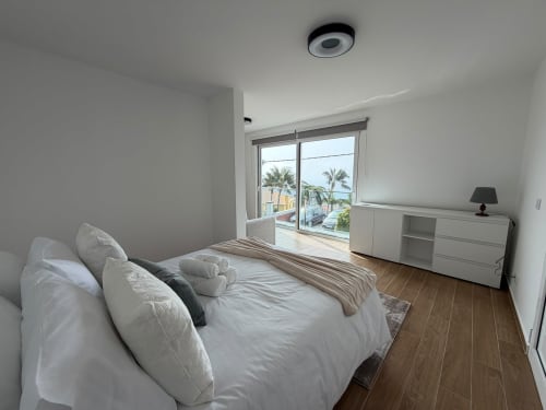 Quarto moderno e luminoso na villa Sunshine 85 House com varanda vista mar, palmeiras visíveis, roupa de cama branca e pisos de madeira