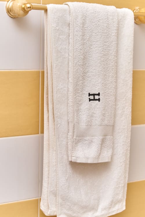 Serviette crème avec monogramme exposée sur porte-serviettes en laiton dans la salle de bain de l'Hôtel Yakro au carrelage jaune et blanc