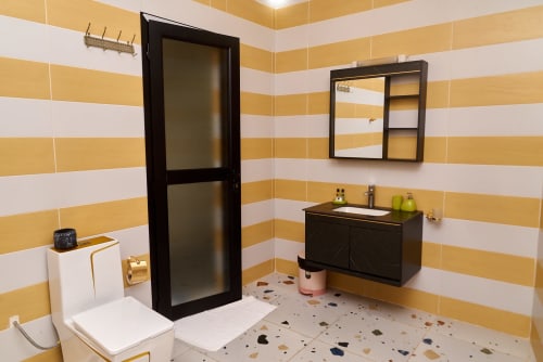 Salle de bain moderne de l'Hôtel Yakro aux murs rayés jaune et blanc, vasque noire, miroir-armoire et douche vitrée