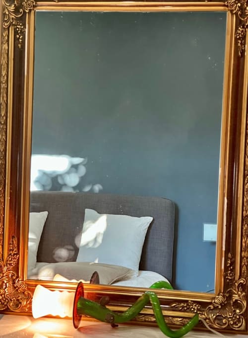 Espelho com moldura dourada ornamentada refletindo quarto com cabeceira cinza e almofadas na Villas Simone