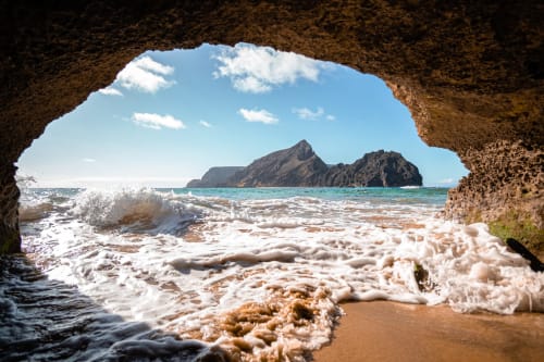 Abertura de gruta rochosa emoldura mar turquesa, praia arenosa com ondas e ilha distante perto de Villas Simone Portugal