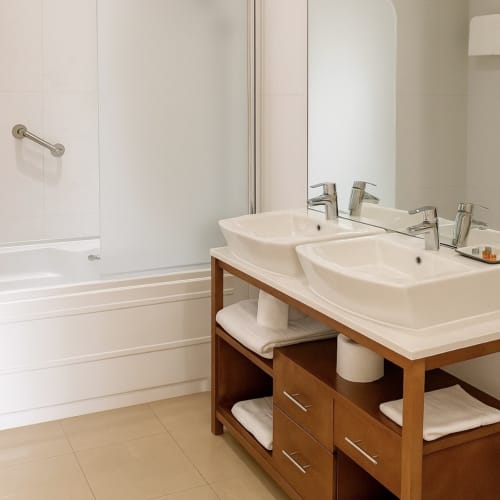 Cuarto de baño con doble tocador moderno en Hotel Villa de Cacabelos, muebles de madera y lavabos blancos de cerámica