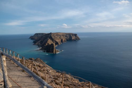 Vista panorâmica de falésia costeira com corrimão de madeira, águas turquesas e ilha ao longe, Portugal