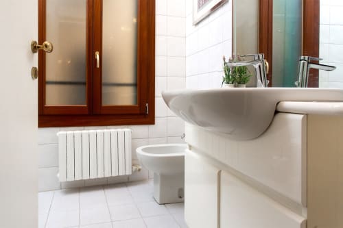Bagno moderno in lussuoso appartamento veneziano con lavabo in ceramica bianca, water, radiatore riscaldato e mobiletto specchio in legno
