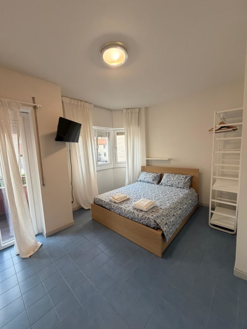 Camera luminosa di Porta Romana Suite Milano con letto in legno, TV, tende bianche e pavimento in piastrelle scure