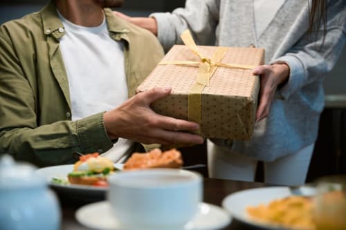 Man presenting wrapped gift at dinner table at Holiday Suites Pesaro with food and plates nearby