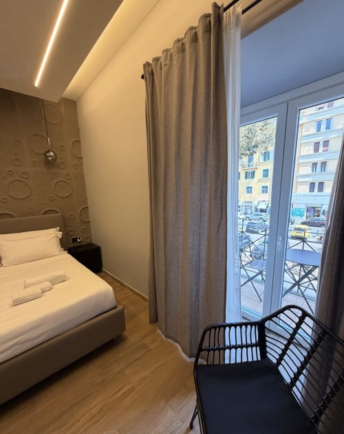 Suite moderna dell'Eterna Suites Roma con letto bianco, tende grigie e balcone con vista sulla strada della città