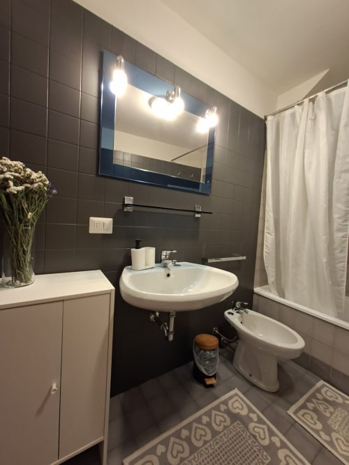 Bagno moderno a Le Arche Verona con piastrelle scure, lavabo rotondo, specchio illuminato, bidet e tenda doccia