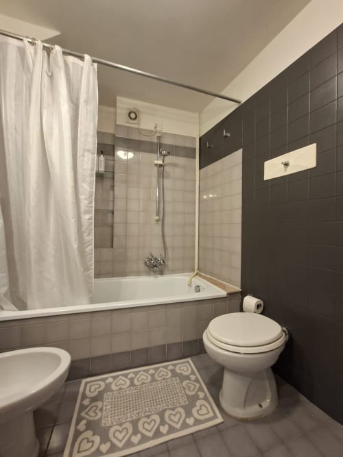 Moderno bagno nell'appartamento Le Arche Verona con vasca, wc, piastrelle scure, sanitari bianchi e tenda doccia