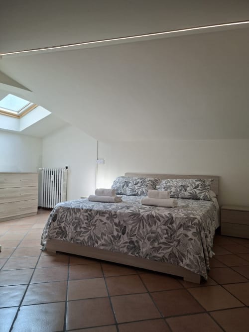 Camera da letto mansardata luminosa con lucernari, letto in legno con biancheria decorata, radiatore bianco e terracotta