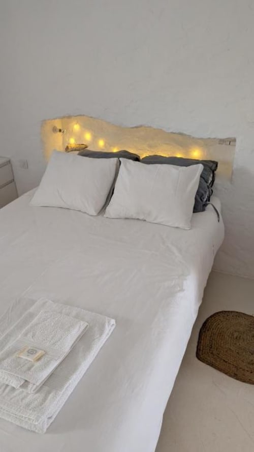Quarto moderno com roupa de cama branca, dois travesseiros brancos e cabeceira ondulada iluminada com luzes quentes em Casas Velhinhas