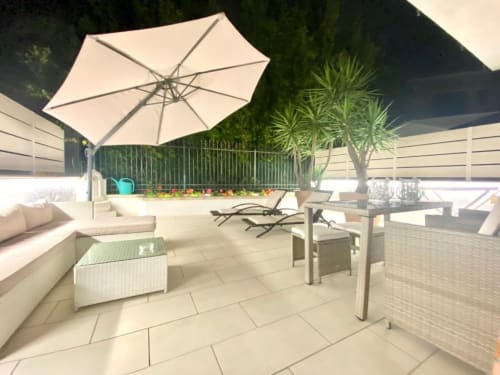 Terrazza soleggiata degli Appartamenti Mottola a Tropea con ombrellone color crema, zona lounge, area pranzo e vista sul giardino