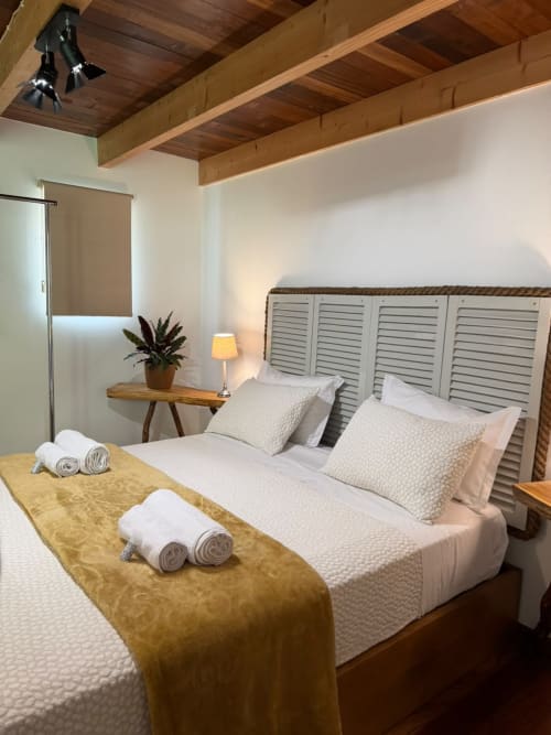 Quarto da hospedaria em Refúgio do Nevoeiro com roupa de cama branca, vigas de madeira, painel cabeceira com venezianas e toalhas enroladas