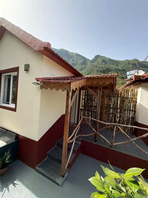 Fachada da cabana no Refúgio do Nevoeiro com pérgula de madeira, pátio com borda vermelha e vistas para a serra em São Vicente, Portugal