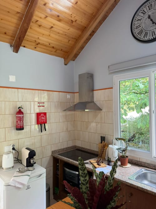 Cozinha rústica com teto de vigas de madeira, azulejos decorativos, fogão com coifa e vista para o jardim no Refúgio do Nevoeiro