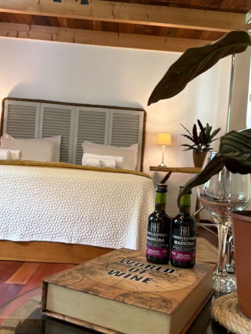 Quarto aconchegante do Refúgio do Nevoeiro com cama, garrafas de vinho Madeira, plantas e luminária de cabeceira
