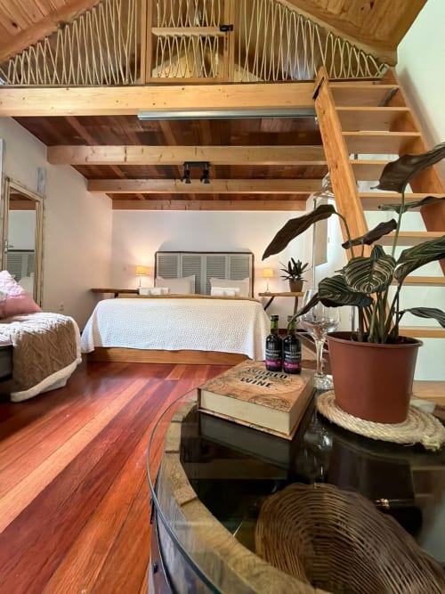 Quarto aconchegante no Refúgio do Nevoeiro com vigas de madeira, cama, planta em vaso, garrafas de vinho e escada de madeira